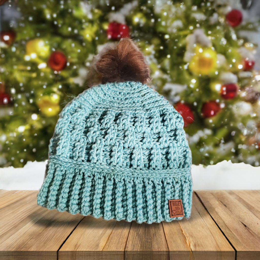 Handmade crocheted aqua winter hat with pompom for women on wooden table.